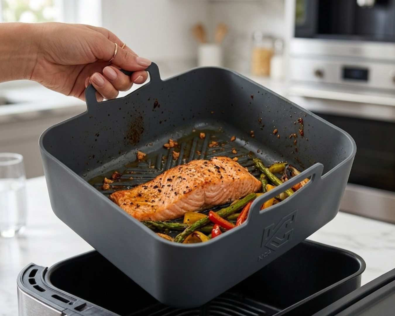 A hand lifts a dark gray square air fryer basket containing cooked salmon and vegetables, showing the release mechanism over a stainless steel air fryer in a bright kitchen setting.