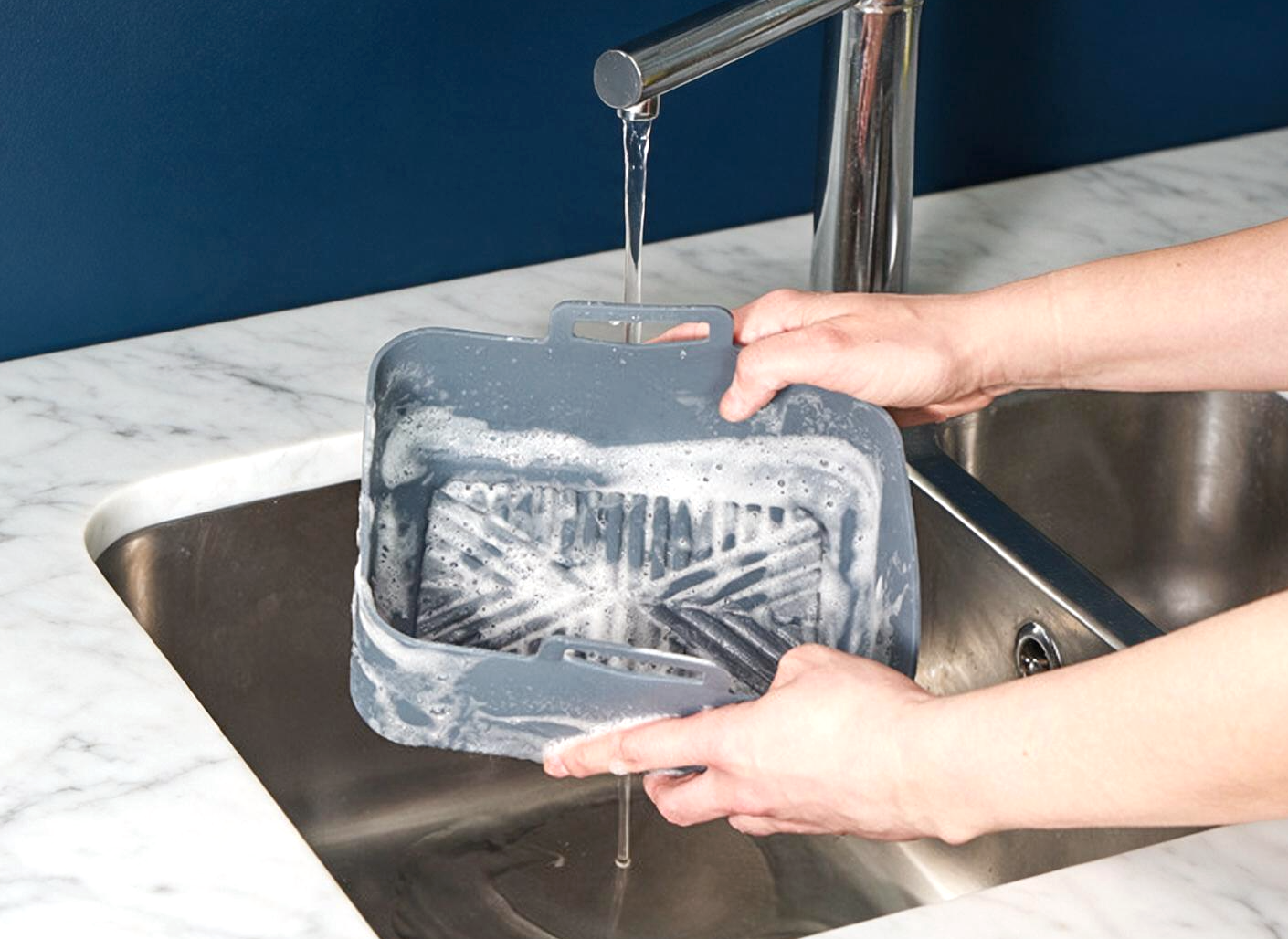 A gray air fryer liner with a handle, covered in soapy foam, being rinsed under running water from a chrome faucet in a kitchen sink.