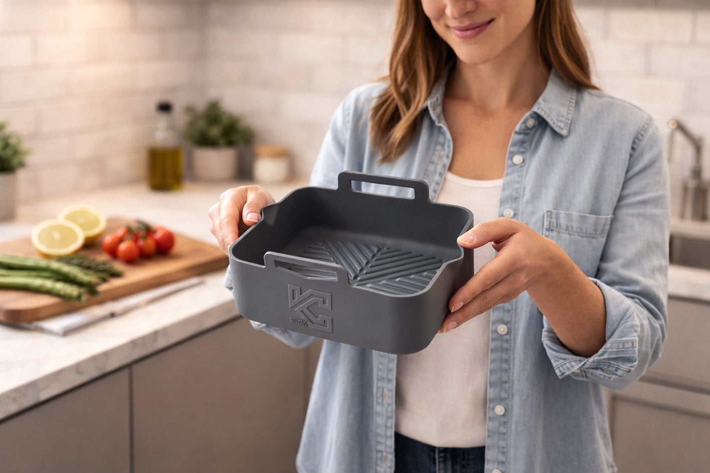 Woman standing in a modern kitchen holding a grey silicone air fryer liner with raised handles and embossed KC logo, with fresh ingredients on the worktop behind her.