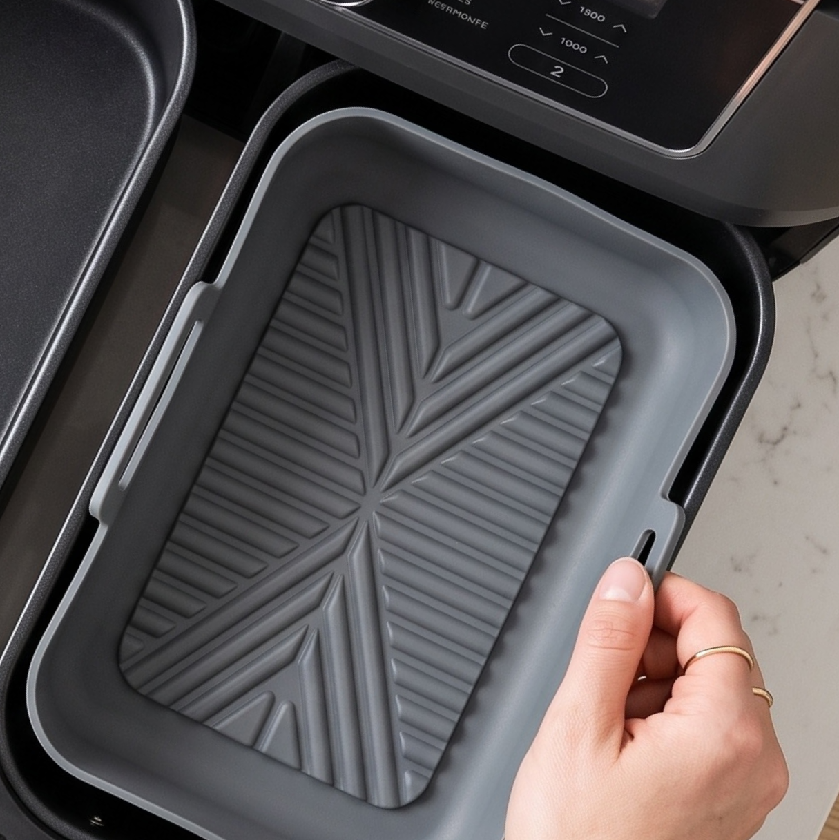 A close-up of a hand inserting a gray rectangular nonstick pan into a black Ninja air fryer basket.