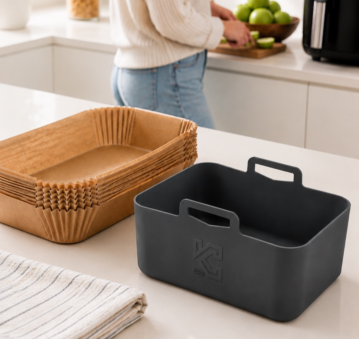 A dark gray silicone storage container with a handle and embossed logo, shown alongside a stack of brown paper liner containers on a kitchen counter, with a person blurred in the background.