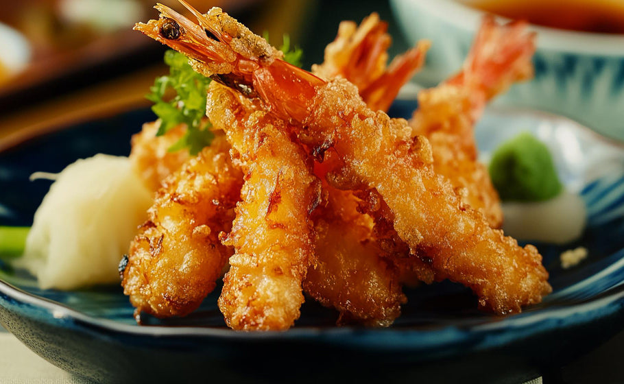 Golden-brown tempura shrimp with crispy battered coating, served on a dark blue plate with small garnishes of green parsley, white radish, and green wasabi in a Japanese restaurant setting.