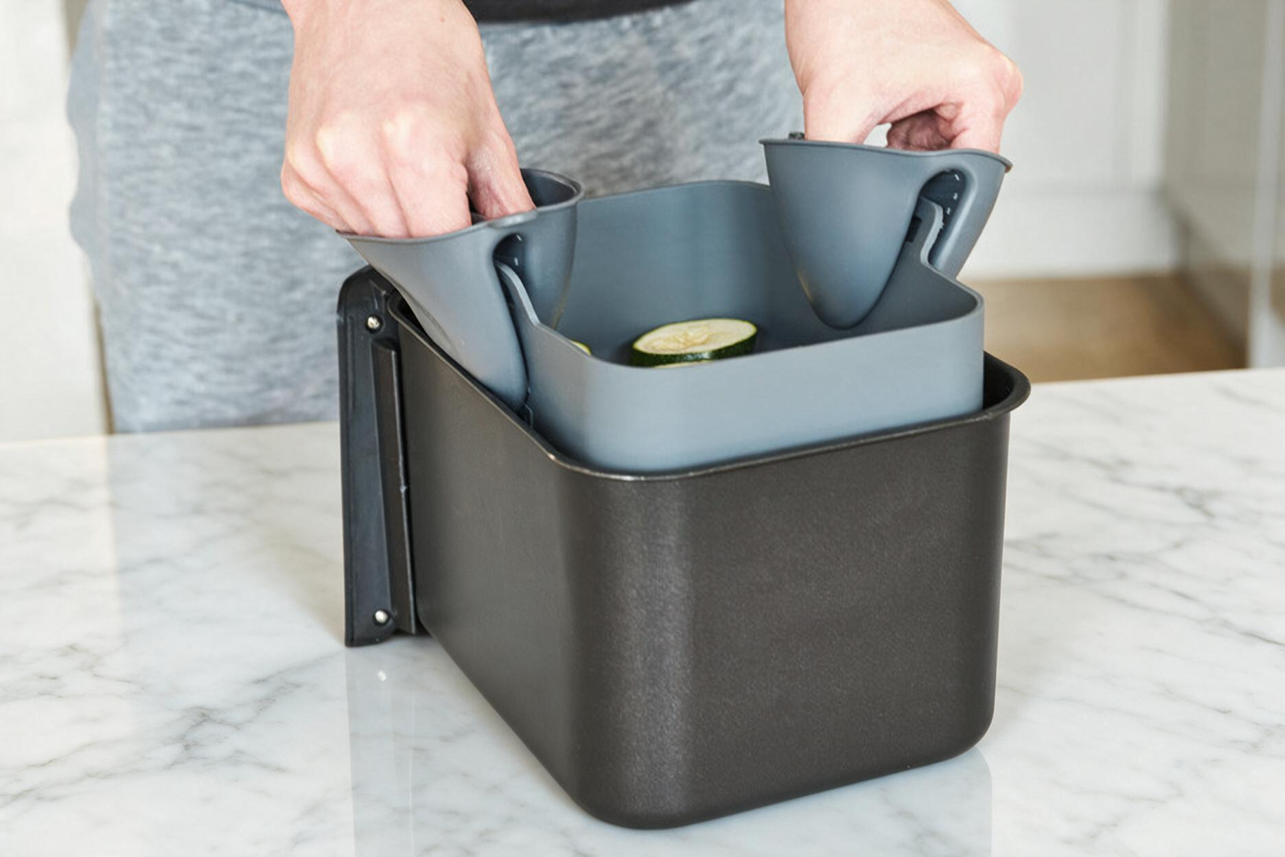 A person's hands are shown taking a platinum air fryer liner, inserting a gray ninja dual  basket mounted on a white marble kitchen counter.